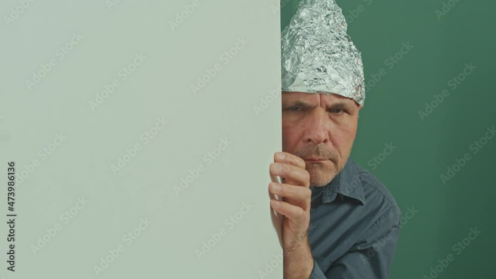 A Frightened Man in a Protective Foil Cap Peeking Strangely Around the Corner, Looking Around Anxiously. Protective Helmet for the Brain from Aliens, 5 G Waves, Electromagnetic Fields. A Frightened Man in a Protective Foil Cap Peeking Strangely Around the Corner, Looking Around Anxiously. Protective Helmet for the Brain from Aliens, 5 G Waves, Electromagnetic Fields.