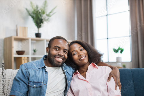 Beautiful and positive young people smiling sincerely on camera while resting together on comfy couch. African american man and woman sitting in hugs at bright living room. © sofiko14