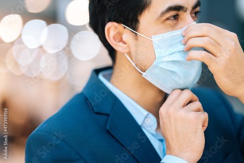 Close-up side view of handsome young man in stylish suit wearing protection face mask looking away in hall of mall centre, blurred background. Closeup of confident male applying mask.