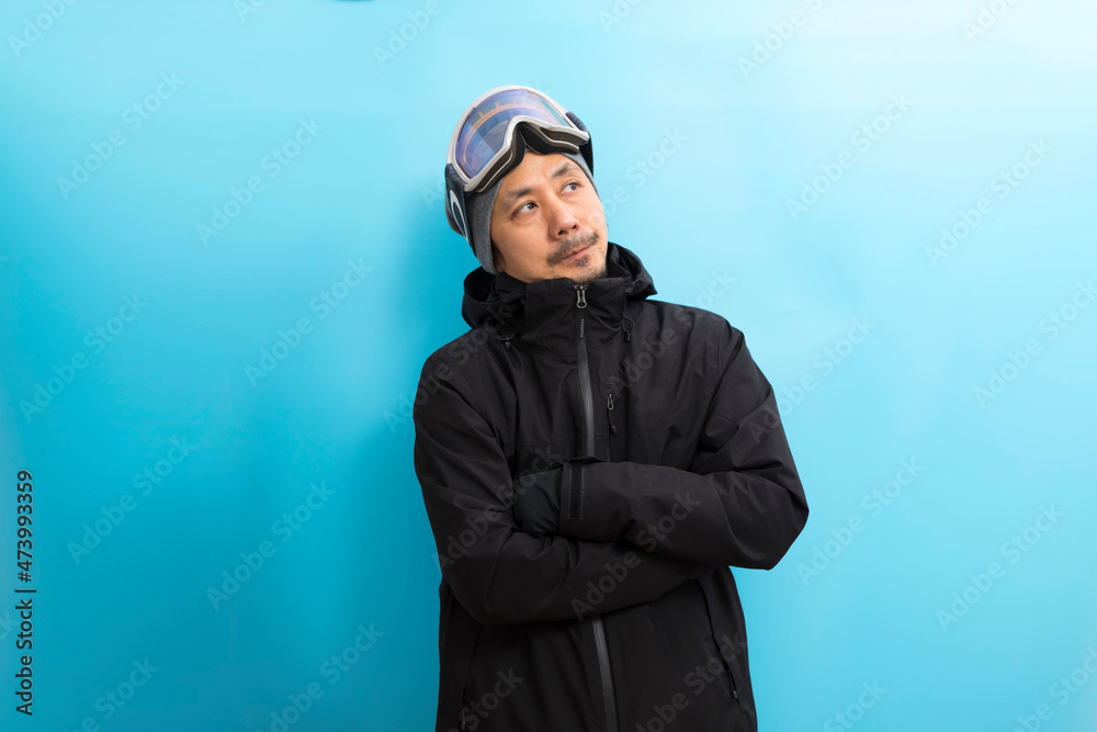 Winter sport. Handsome asian man with snowboard goggles and black anorak coat with curious face stand on blue background. Winter sport. Handsome asian man with snowboard goggles and black anorak coat with curious face stand on blue background.