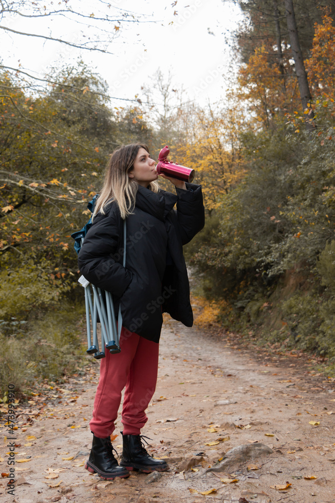 Traveler girl having rest and hot drink from thermos in a nature park on a gold autumn day. She has chair in her back. Young nice looking blonde girl admire autumn nature landscape. Traveler girl having rest and hot drink from thermos in a nature park on a gold autumn day. She has chair in her back. Young nice looking blonde girl admire autumn nature landscape.