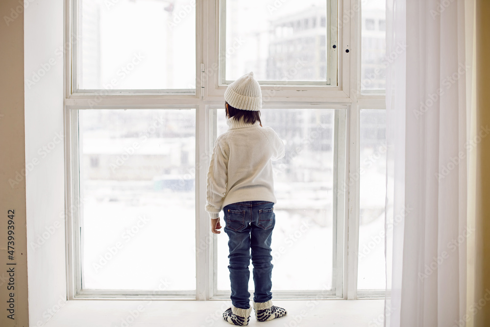 boy in a white sweater and hat stands at the window in winter in large apartment on Christmas day