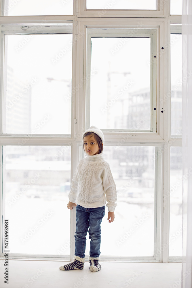 boy in a white sweater and hat stands at the window in winter in large apartment on Christmas day boy in a white sweater and hat stands at the window in winter in large apartment on Christmas day