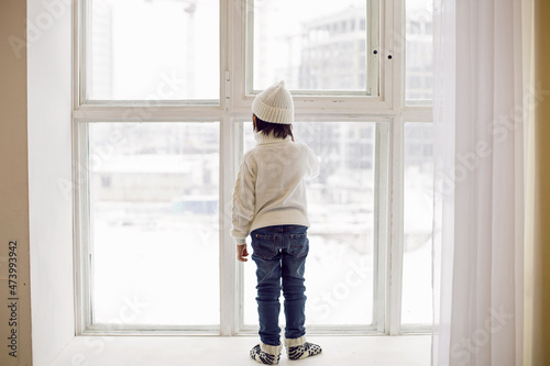 Obraz na płótnie boy in a white sweater and hat stands at the window in winter in large apartment on Christmas day w salonie