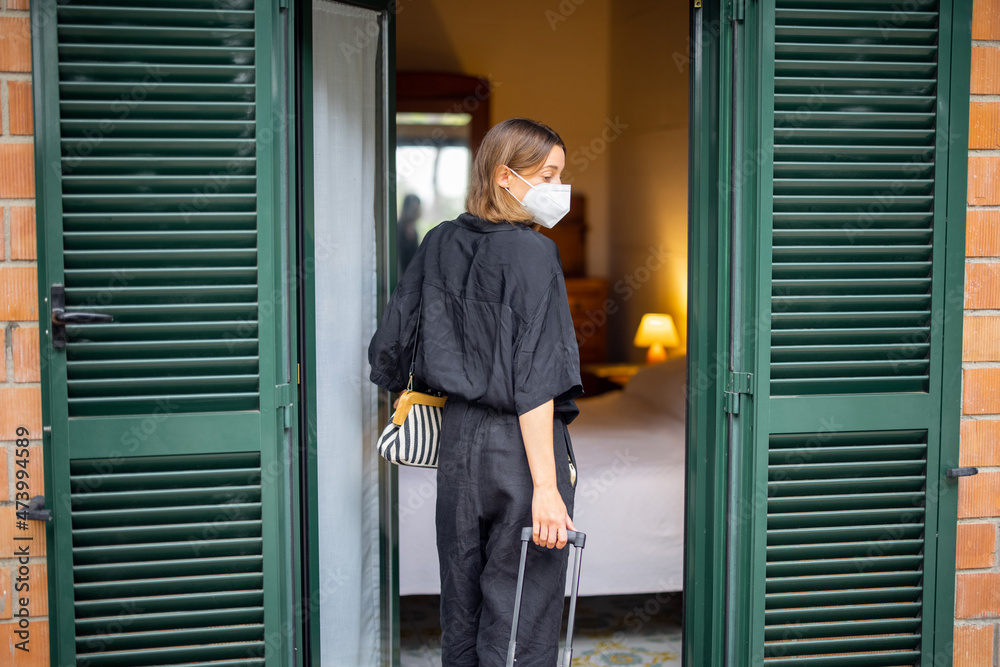 Woman in medical mask with baggage entering in hotel room. Concept of weekend, rest, tourism and vacation. Idea of health protection during Coronavirus pandemic. Young caucasian girl Woman in medical mask with baggage entering in hotel room. Concept of weekend, rest, tourism and vacation. Idea of health protection during Coronavirus pandemic. Young caucasian girl