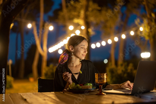 Caucasian woman watching something on laptop computer at evening time outdoors. Young woman sitting at table, eating fruits and drinking wine. Concept of weekend, rest and vacation. Idea of leisure