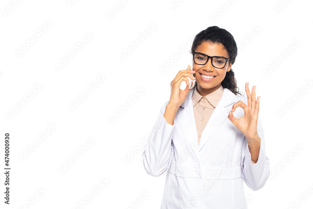Smiling african american ophthalmologist talking on smartphone and showing ok sign isolated on white. Smiling african american ophthalmologist talking on smartphone and showing ok sign isolated on white.