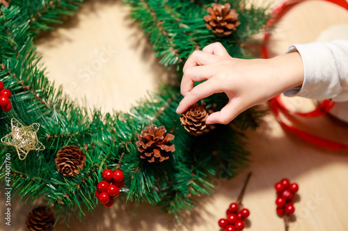 Child's right hand putting pine cone to decorate Christmas wreath on wooden background with decorative red ribbon, golden stars and red berries.