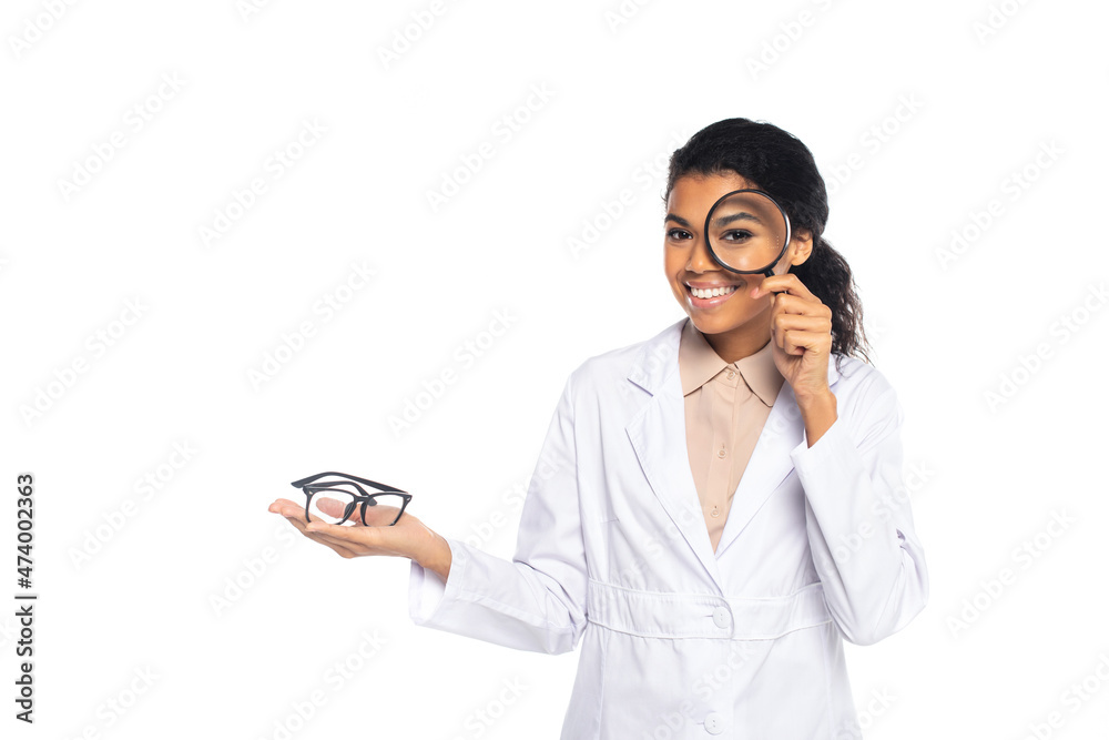 Smiling african american oculist holding eyeglasses and magnifying glass isolated on white. Smiling african american oculist holding eyeglasses and magnifying glass isolated on white.
