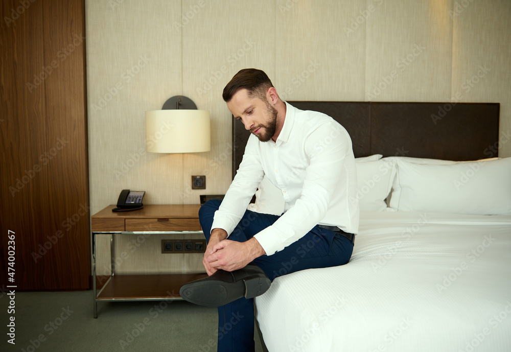 Attractive Caucasian middle aged man, successful businessman on a business trip, sits on the edge of the bed in a hotel bedroom and ties the laces on his shoes
