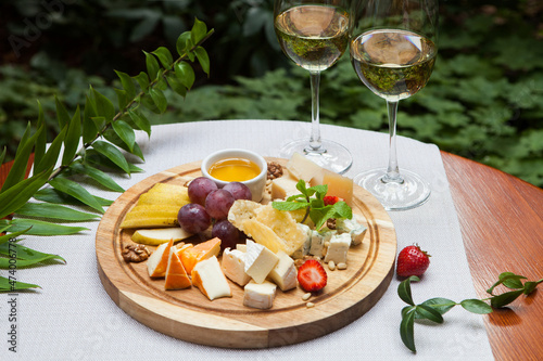 Cheese plate  sliced cheese of different kinds  honey  and fruit on a wooden tray. Next to the glasses with white wine.