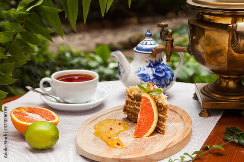 A piece of honey cake, on a wooden plate with fruit. A cup of tea and a samovar in the background.