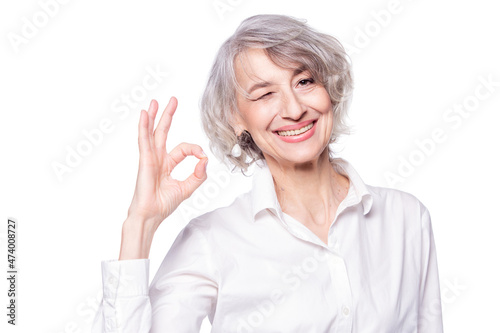 Close up portrait of mature attractive woman wearing elegant shirt standing over isolated white background smiling with happy face winking at camera doing okay sign
