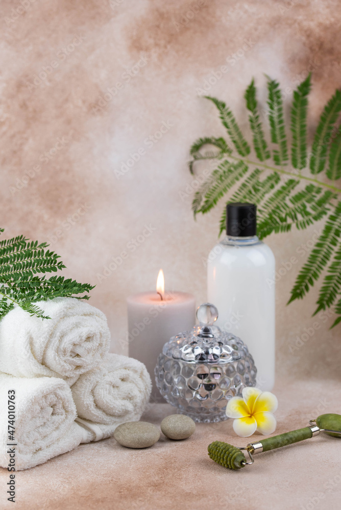 Spa still life treatment composition on massage table in wellness center. Twisted hot towel with aromatic candles and face roller gua sha on beige background. Aroma therapy setting. 
