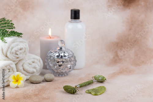 Spa still life treatment composition on massage table in wellness center. Twisted hot towel with aromatic candles and face roller gua sha on beige background. Aroma therapy setting. 