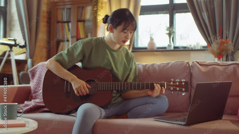 Young Asian woman sitting on sofa at home, watching video lesson on laptop and learning how to play guitar