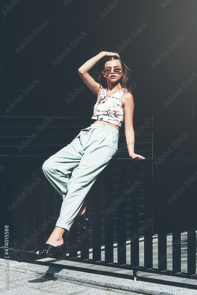 Fashion portrait of young woman wearing sunglasses, top, slingbacks, blue suit. Young beautiful happy model posing near gray metal grid