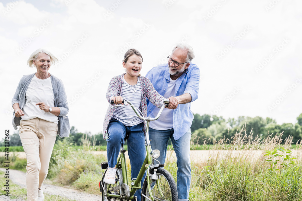 Man teaching girl to ride bicycle with woman running behind on dirt road Man teaching girl to ride bicycle with woman running behind on dirt road