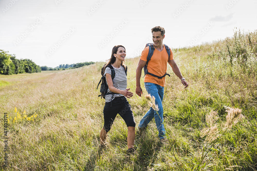 Smiling couple with backpacks hiking together on grass during sunny day Smiling couple with backpacks hiking together on grass during sunny day