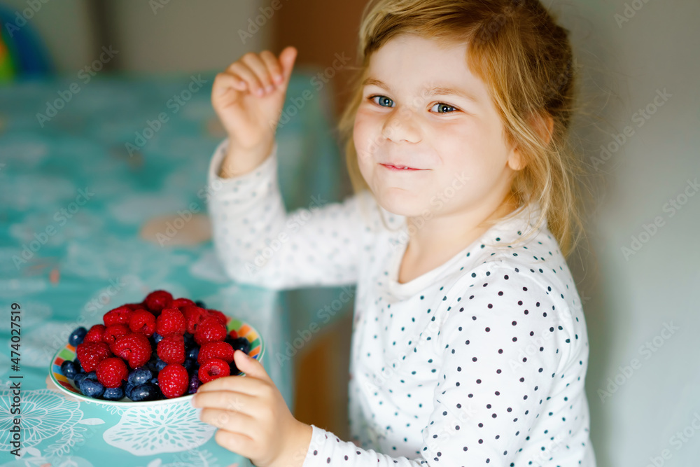 Cute little preschool girl eating fresh raspberries and blueberries. Happy child tasting raspberry and blueberry. Healthy food, childhood and development. Happy kid at home or nursery. Cute little preschool girl eating fresh raspberries and blueberries. Happy child tasting raspberry and blueberry. Healthy food, childhood and development. Happy kid at home or nursery.