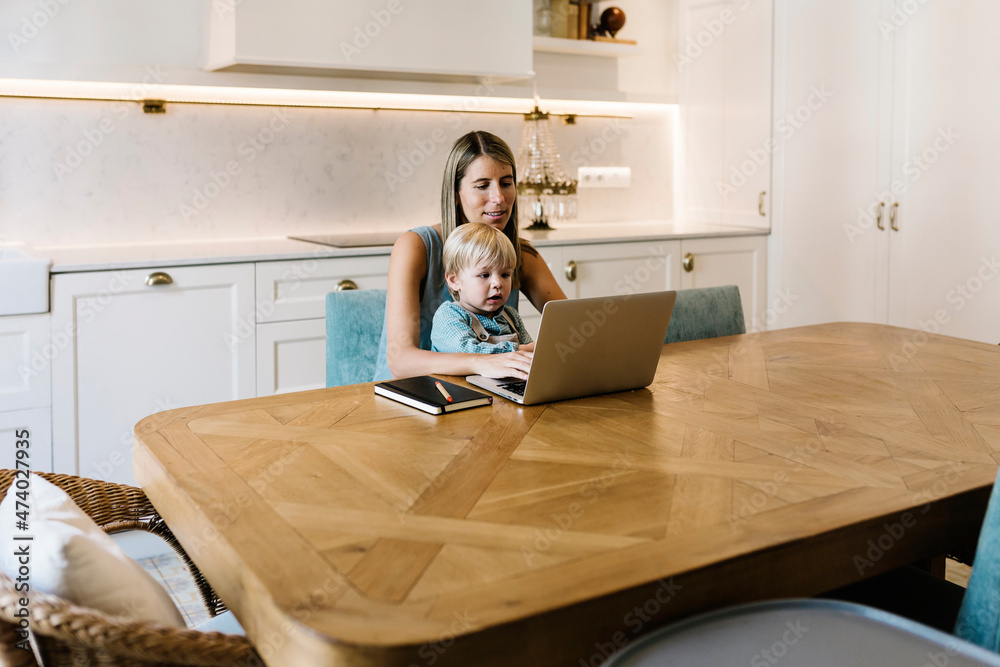 Businesswoman with son working on laptop at dining table in kitchen Businesswoman with son working on laptop at dining table in kitchen