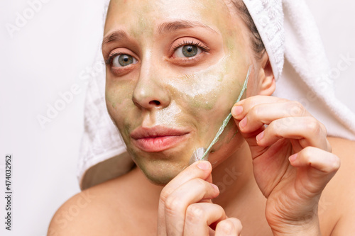 A young beautiful caucasian woman with a white towel on her head after a shower removing a green peeling mask with glitter for the face on a white background. Skin care, cosmetology