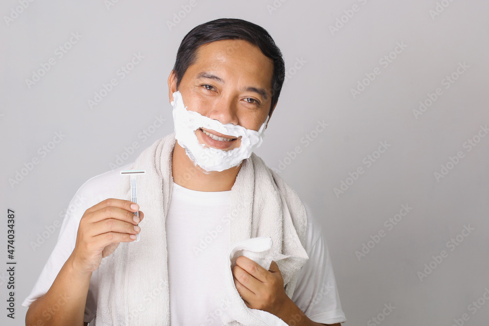 Handsome asian man shaving. Handsome asian man shaving his face and looking at camera while standing on grey background