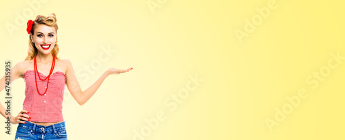 Wide photo of happy excited smiling woman holding, giving, promote, advertising something. Blond girl in pinup style, showing copy space. Retro fashion and vintage. Light yellow background