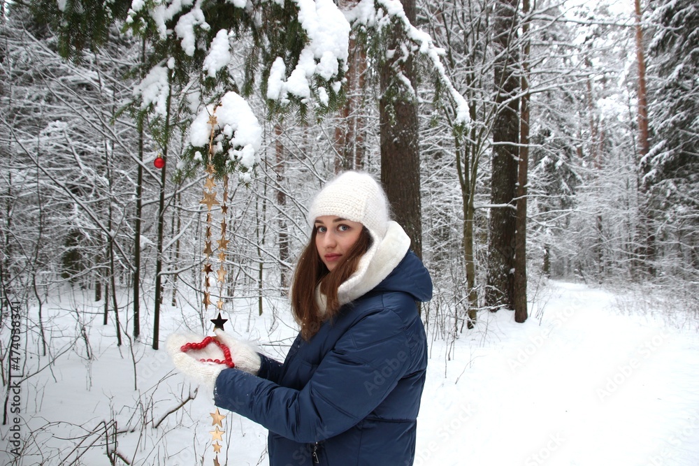 Young attractive woman with long hair, blue eyes, dressed in blue coat, white hat, mittens holds red beads in heart form  in beautiful winter forest among snowy trees