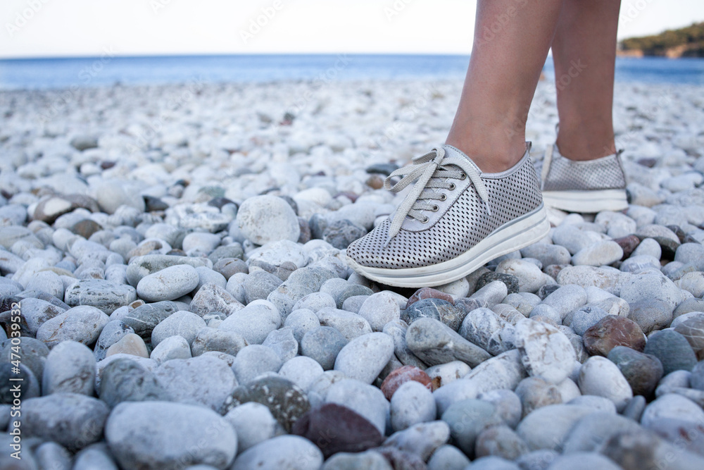 Silver leather sneakers shoe on a female leg walking on beach. Walking on  pebble beach in comfortable shoes. Stock Photo | Adobe Stock