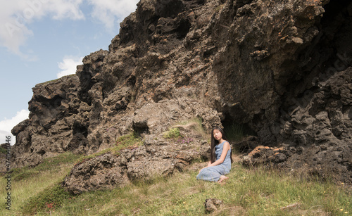 girl in blue dress sitting near wall of black lava rocks in iceland in summer
