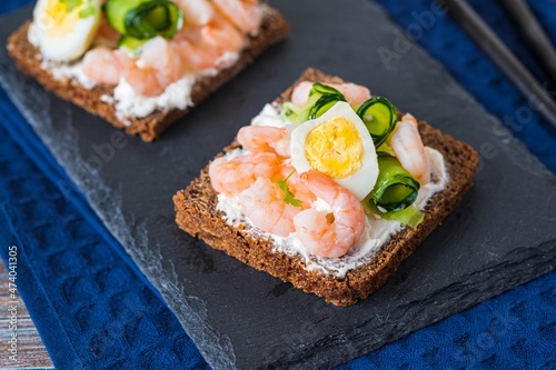 Smorrebrod, open sandwich with shrimps, creme cheese, boiled quail egg and cucumber slices on a slate or stone board on a wooden background. Sandwich recipes.