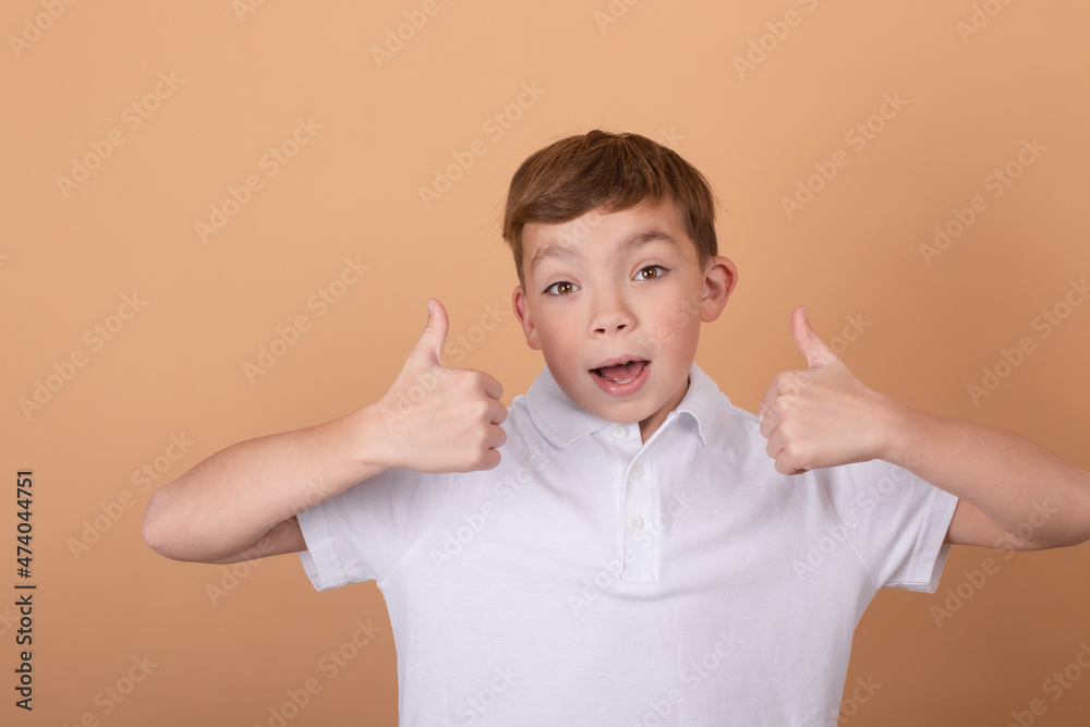 Things are good. Pretty school boy in white t-shirt looking at the camera and showing thumbs up gesture on light brown background. Portrait of handsome teenager guy 9-11 years old with like sign. Things are good. Pretty school boy in white t-shirt looking at the camera and showing thumbs up gesture on light brown background. Portrait of handsome teenager guy 9-11 years old with like sign.