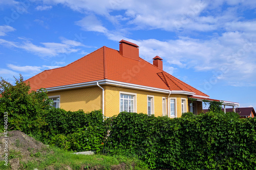 Yellow suburban brick house red roof with a fence of dense green vegetation in bright sunny summer