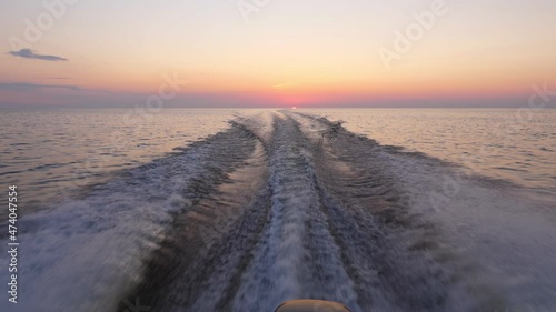 Sailing at calm Ladoga lake in sunset, thin stripe of sun seen at horizon, clear sky in beautiful yellow and blue shades. Look back from rushing vessel, waves from motor split flat surface of water photo