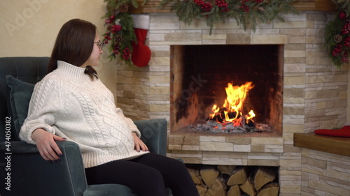 A young pregnant woman with glasses in a sweater sits in an armchair by the fireplace and looks at the fire. Christmas mood.