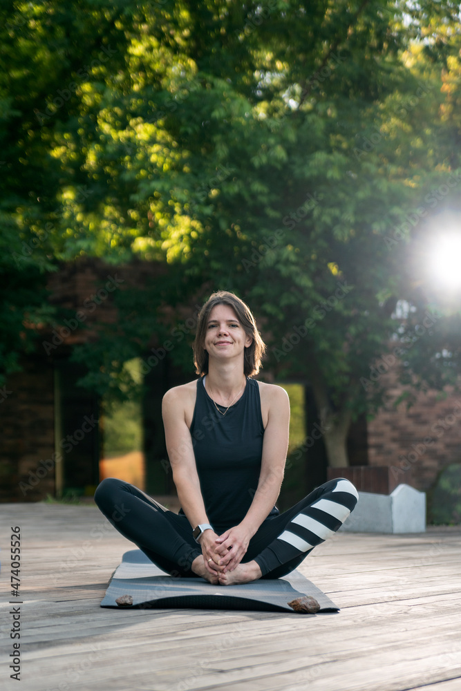 Portrait of young woman in sportswear sits on yoga mat outside. Morning yoga practice outdoors. Portrait of young woman in sportswear sits on yoga mat outside. Morning yoga practice outdoors.
