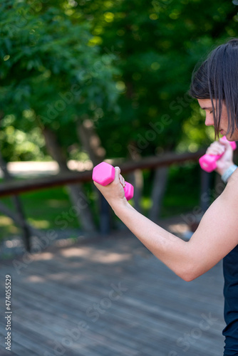 Sports girl holds small womens pink dumbbells. Young woman does outdoor fitness. Copy space. Vertical frame