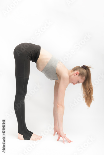Young woman in tight leggings and top is doing stretching. Girl reaches for the floor with her hands on white background. Exercise for flexibility.