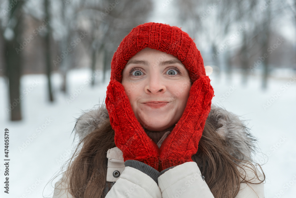 Winter portrait of cheerful girl in red knitted hat and mittens on snow-covered forest background. Girl warms her cheeks with her hands in gloves Winter portrait of cheerful girl in red knitted hat and mittens on snow-covered forest background. Girl warms her cheeks with her hands in gloves