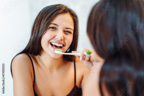 Young beautiful woman brushes her teeth in the morning in the bath looking into her reflection in the mirror