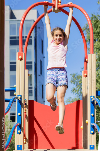 A positive smiling girl spends time on the playground of the yard, hanging on the crossbar of a sports equipment. 