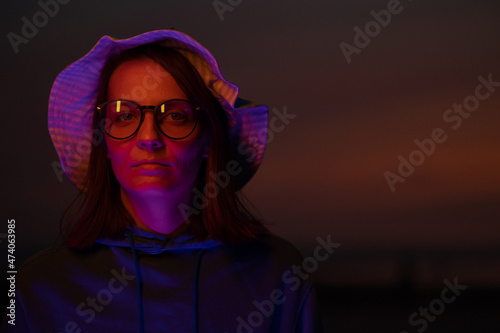 a European woman illuminated by neon light in a hat against the background of the sunset sky. neon lights and a woman at sunset. stylish cyber photo. color filters on neon lamps. creative photo and