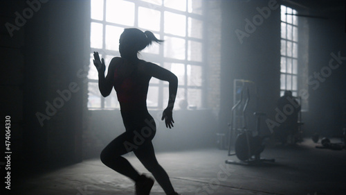 Active sportswoman jogging in dark loft building. Strong woman running in gym