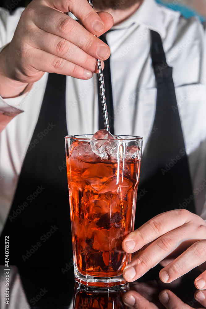 The hand of a professional bartender stirs the red syrup in an alcoholic cocktail with a bar spoon on the bar counter. The process of preparing an alcoholic beverage The hand of a professional bartender stirs the red syrup in an alcoholic cocktail with a bar spoon on the bar counter. The process of preparing an alcoholic beverage
