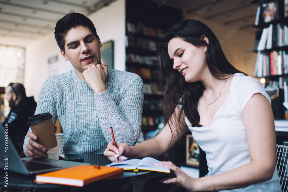 Young female student taking notes in copybook while preparing for exam with friend
