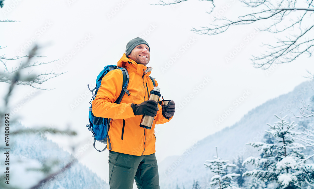 Smiling Man under snowfall dressed bright orange softshell jacket with a hot drink thermos flask while he trekking winter mountains route. Active people in the nature concept image.