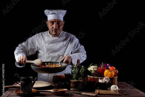 Male cook in white uniform and hat with ladle mixes the ingredients onto the cooking pan photo