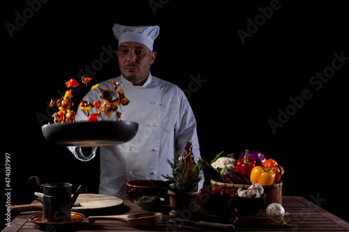 Male chef in white uniform holding a frying pan, sautéing fresh vegetables flying in the air photo