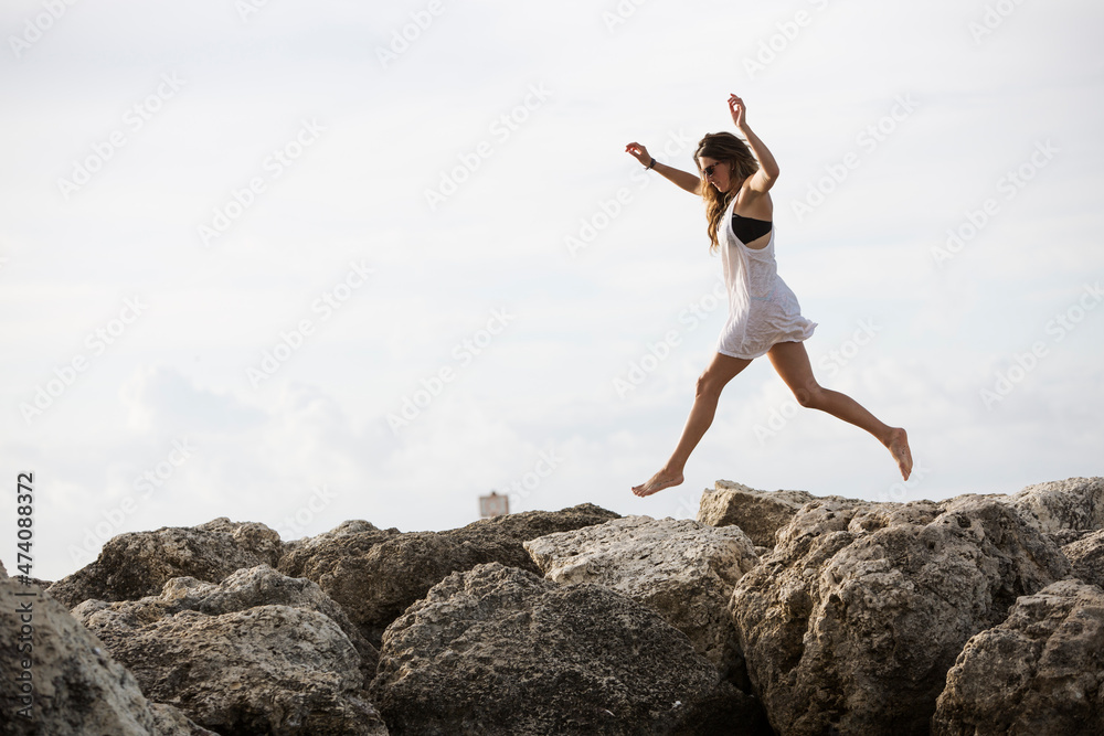 An attractive young woman jumps mid air along shoreline rocks An attractive young woman jumps mid air along shoreline rocks